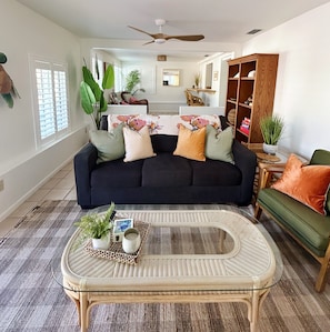 A living room with a dark blue sofa, a wicker coffee table, and a green armchair. There is a wooden bookshelf and a ceiling fan. - Poolside Palms & Oak; heated saltwater pool, 10 min to beach, A/C, dog-friendly (Venice)