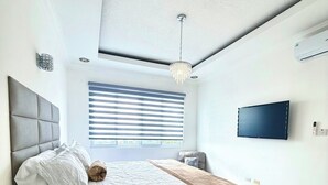 Ein Schlafzimmer mit Bett, Nachttisch, Fernbedienungen, Fernseher und einem Fenster mit Jalousien.