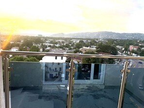 Ein Balkon mit Glasgeländer mit Blick auf eine Stadtlandschaft, Gebäude und eine Bergkette in der Ferne.