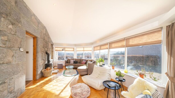 A cozy living room with a stone wall, a fireplace, a sofa, and large windows offering a view of the countryside. - Killiegowan Self-catering Holiday Home in Aberdeen (Aberdeen)