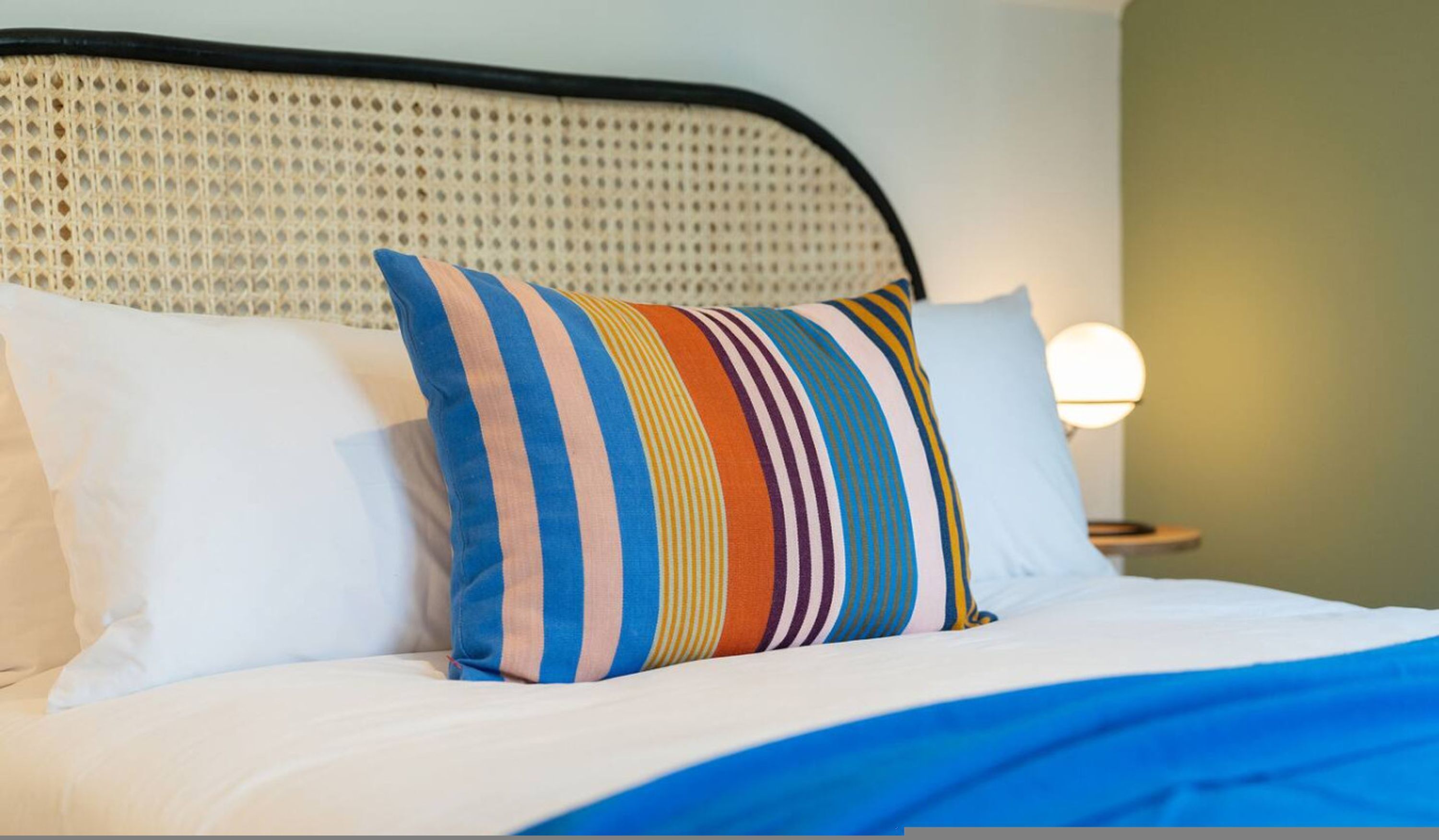 A bed with white bedding and a colorful striped pillow, a woven headboard, and a bedside lamp.