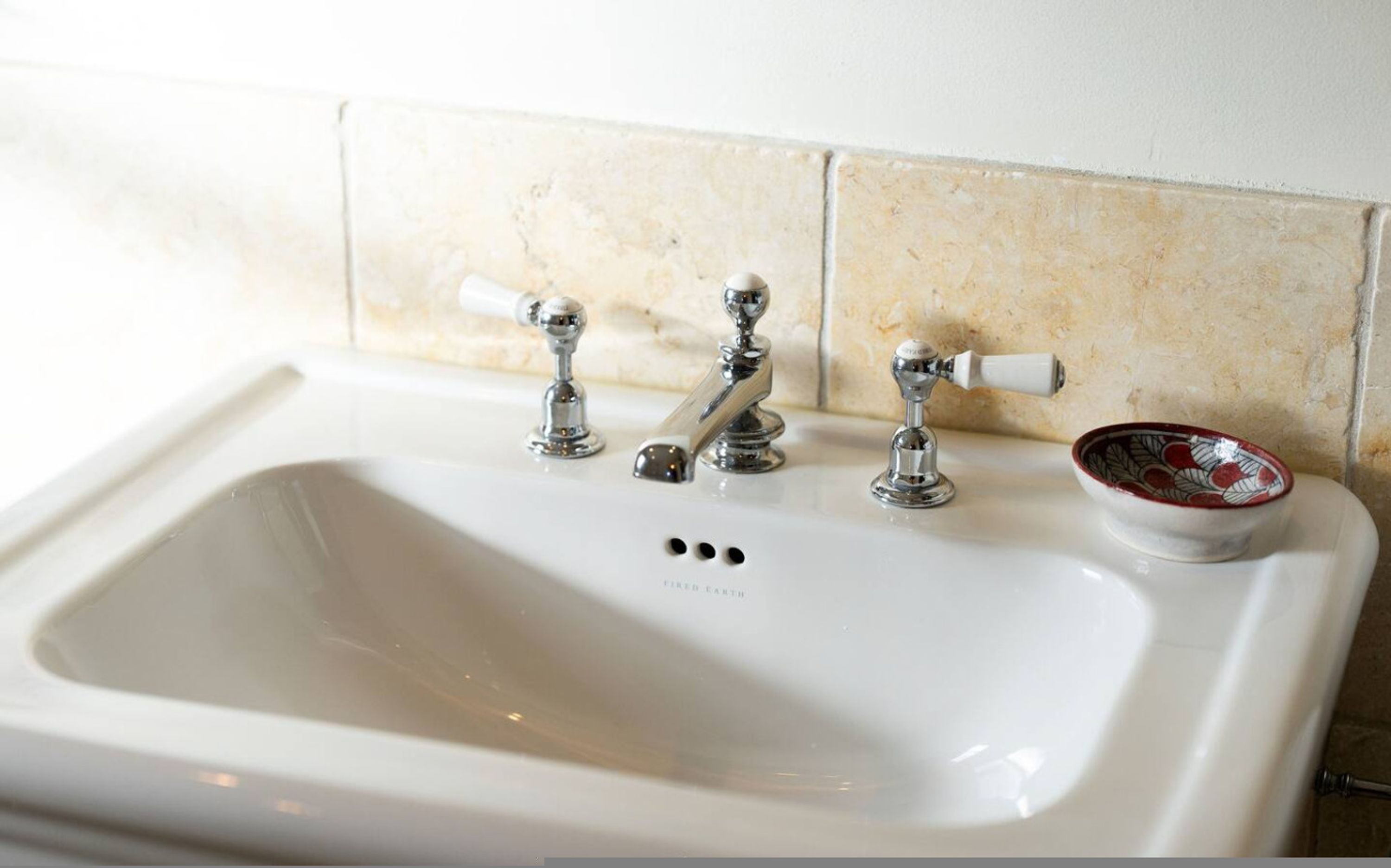 A bathroom sink with three faucets, a white basin, and a decorative bowl with a floral pattern.