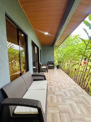 Un área de estar al aire libre techada con muebles de mimbre, piso de baldosas y plantas en macetas.
