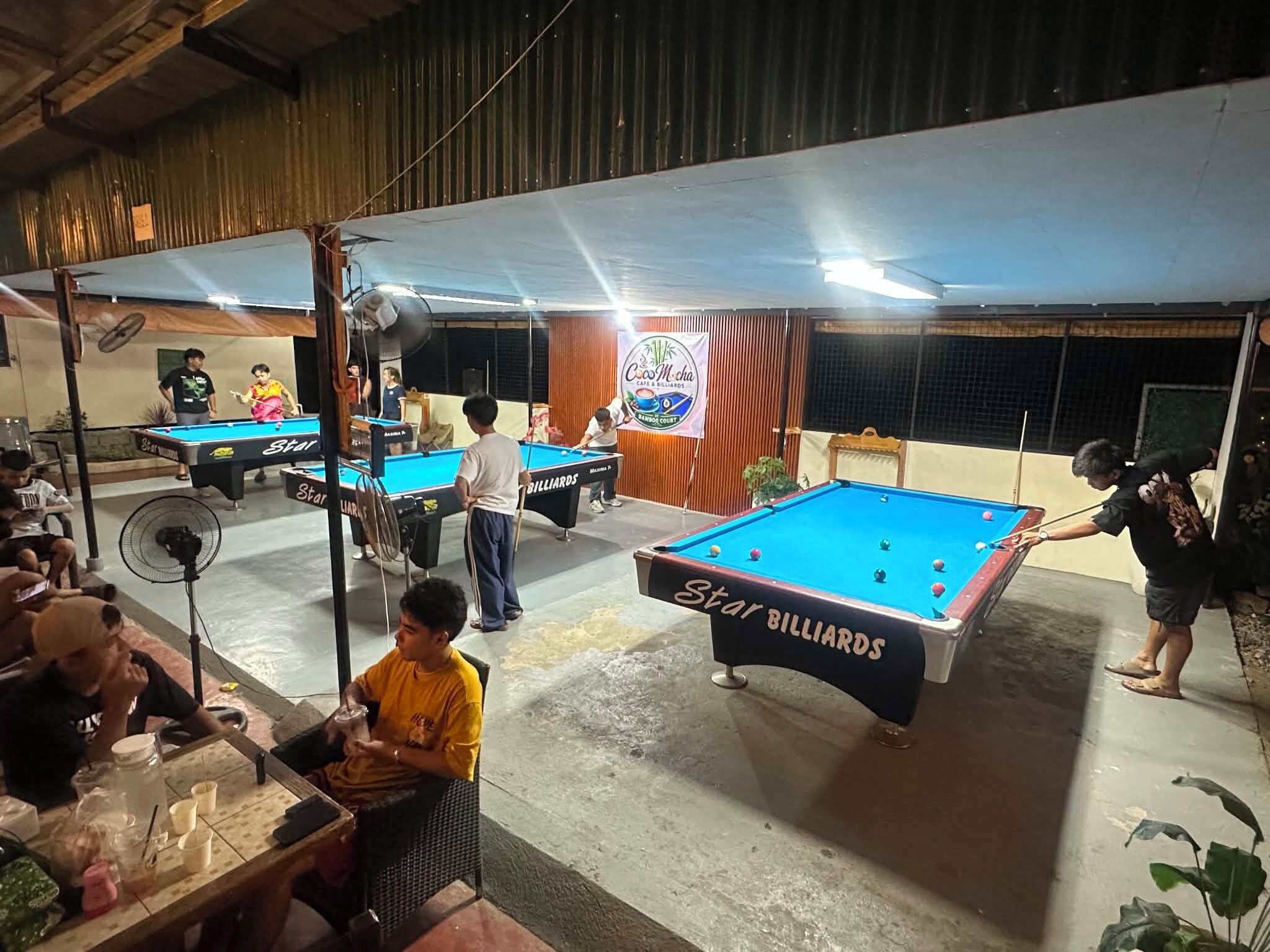 Una sala de billar amb diverses taules de bitlles, un pancartes de cortina de bambú i una zona de seients informal amb begudes.