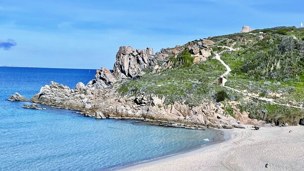 Paisagem costeira com falésias rochosas, mar azul cristalino e um caminho que sobe a colina.