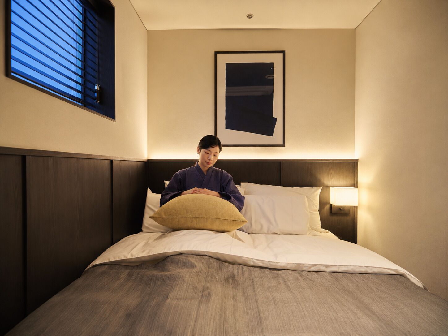 A person sitting on a bed with a pillow, a framed picture on the wall, and a window with blinds.