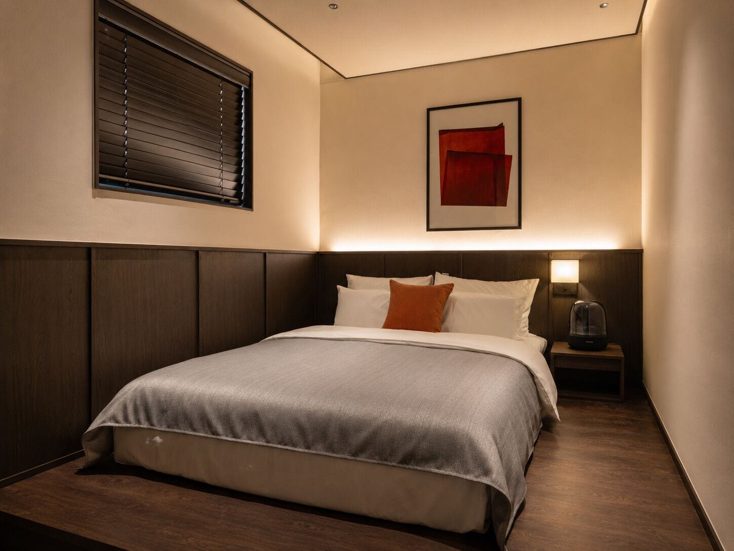 A hotel room with a bed, a nightstand, a lamp, a framed picture on the wall, and a window with blinds.