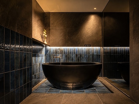 A modern bathroom with a large freestanding bathtub, dark tiled walls, and a small vase with flowers.