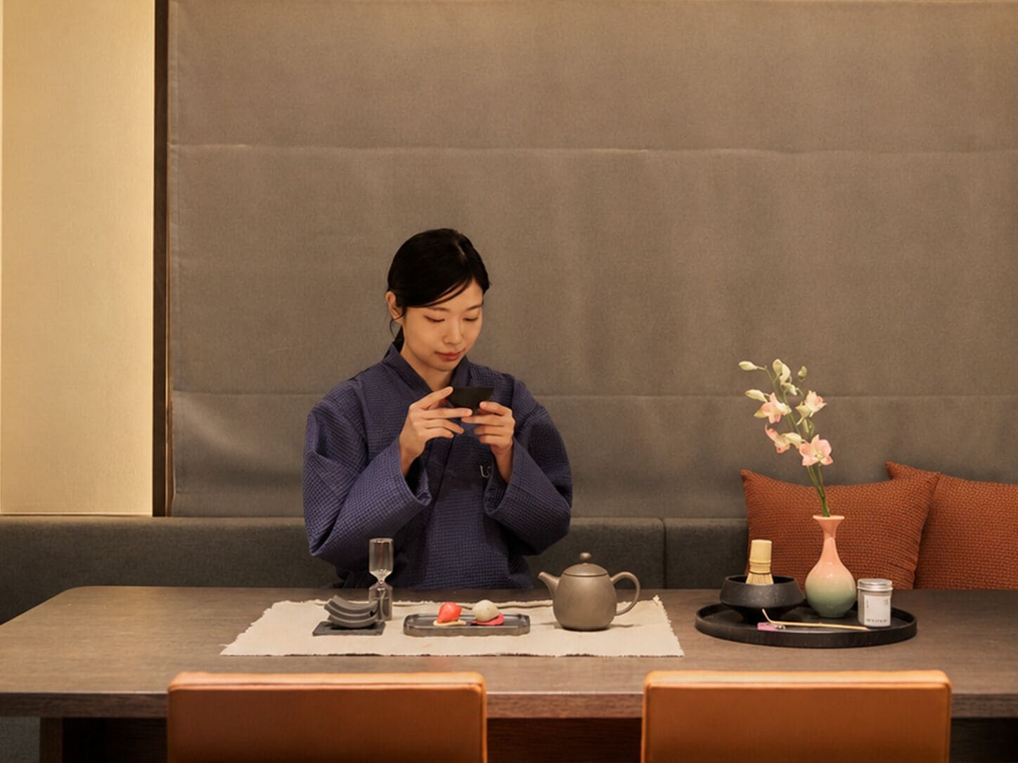 A person seated at a table with a teapot, a vase with flowers, and a tray with items.
