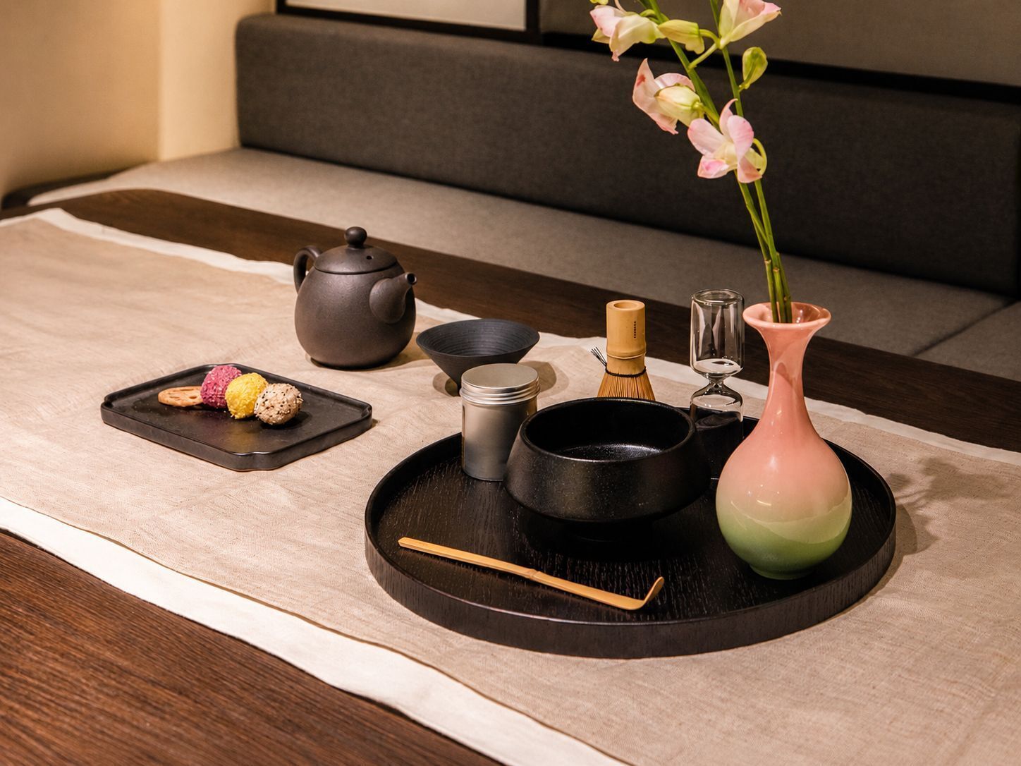 A traditional Japanese tea setting with a teapot, cups, a bamboo whisk, a vase with flowers, and a tray with sweets.