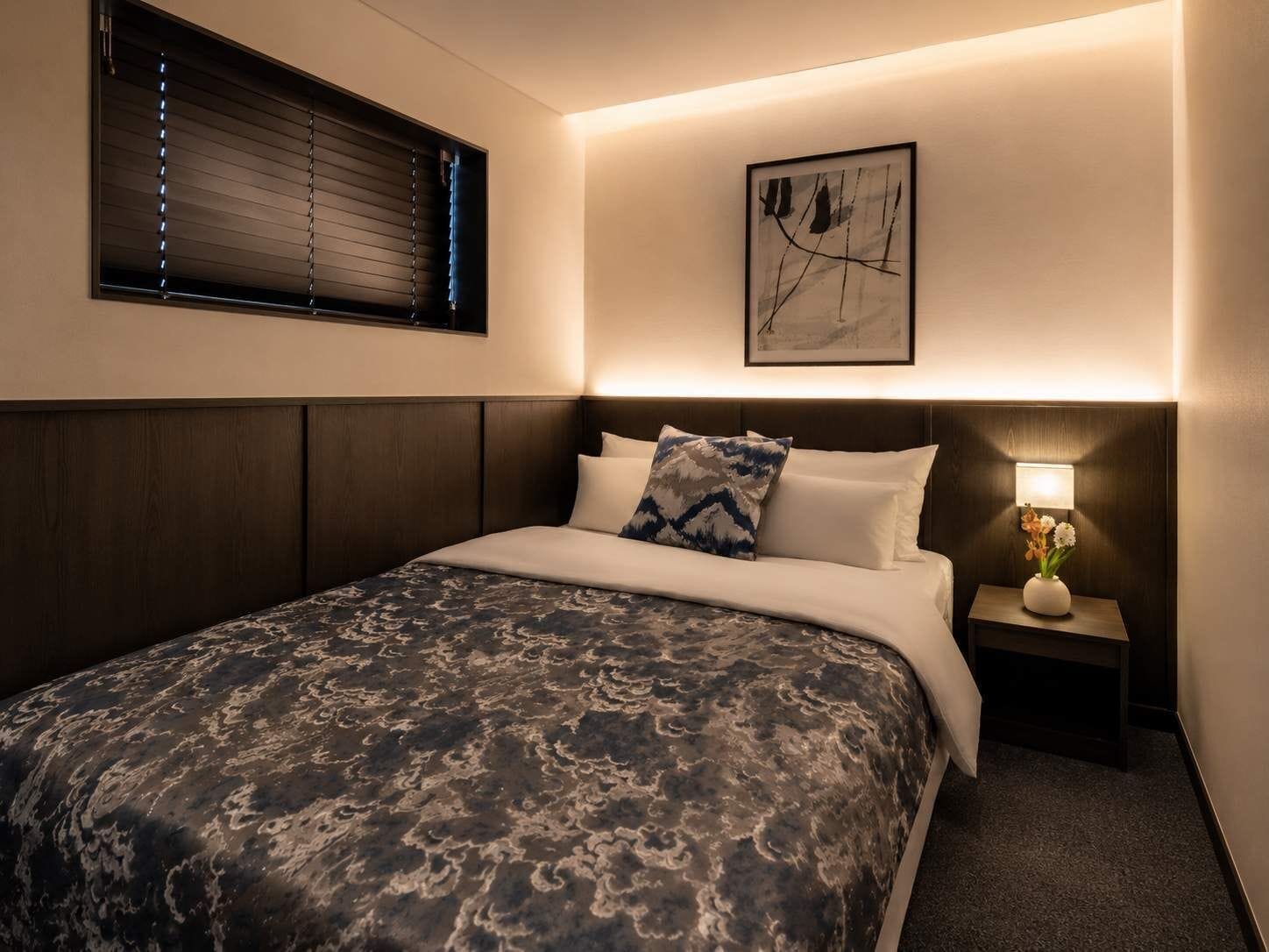 A hotel room with a bed, a nightstand with a lamp, a painting on the wall, and a window with blinds.