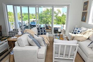 A spacious living room with a large window overlooking a pool and lush greenery. There are multiple seating areas with white and blue furnishings.