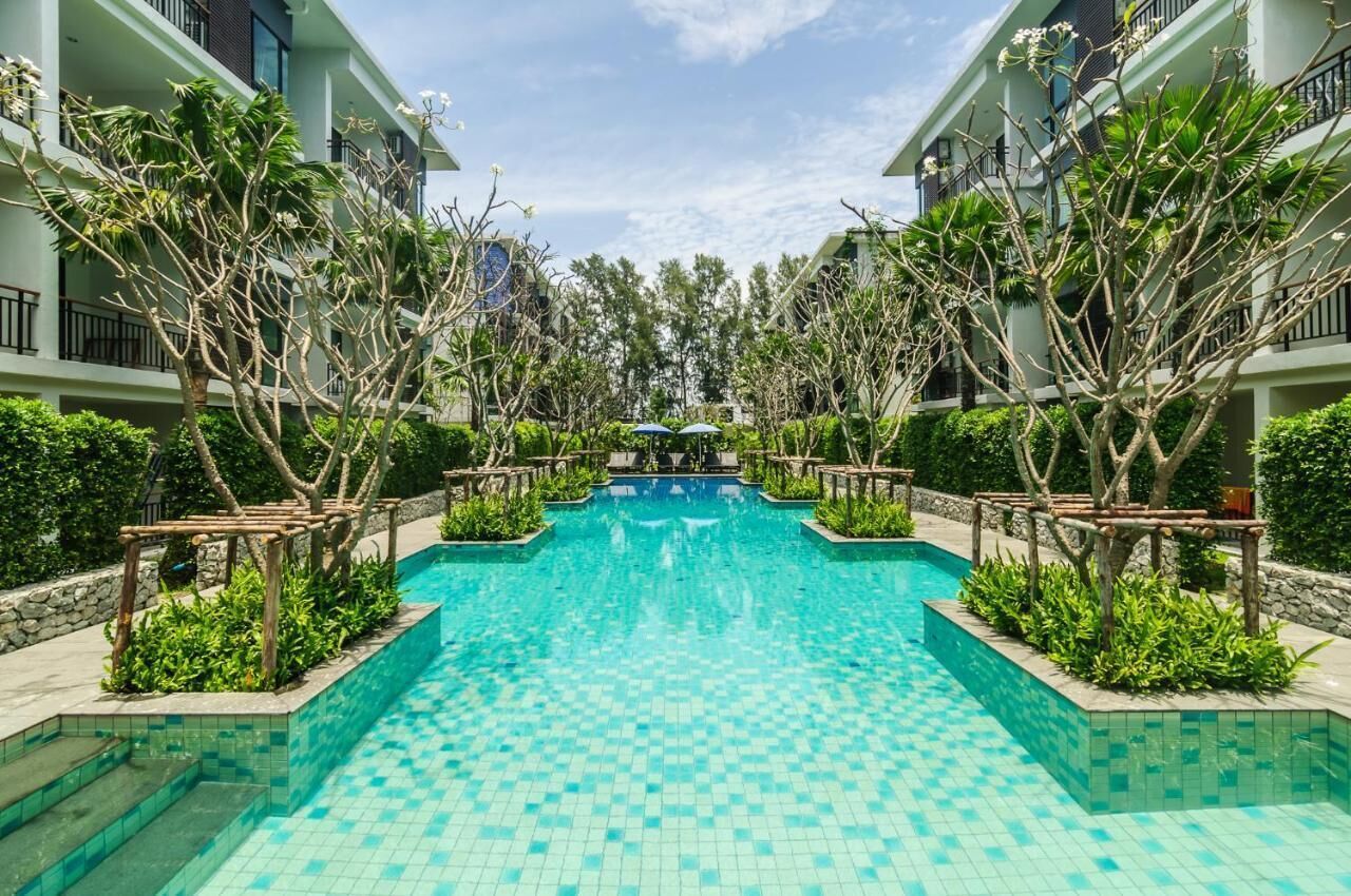 A swimming pool with a tiled floor, surrounded by trees and plants, and a building with balconies in the background.