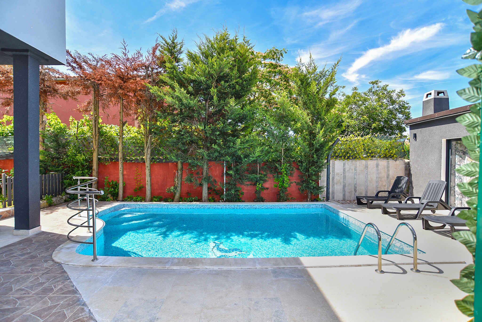A rectangular swimming pool with a tiled edge, surrounded by a concrete deck. There are lounge chairs and a small structure with a chimney.