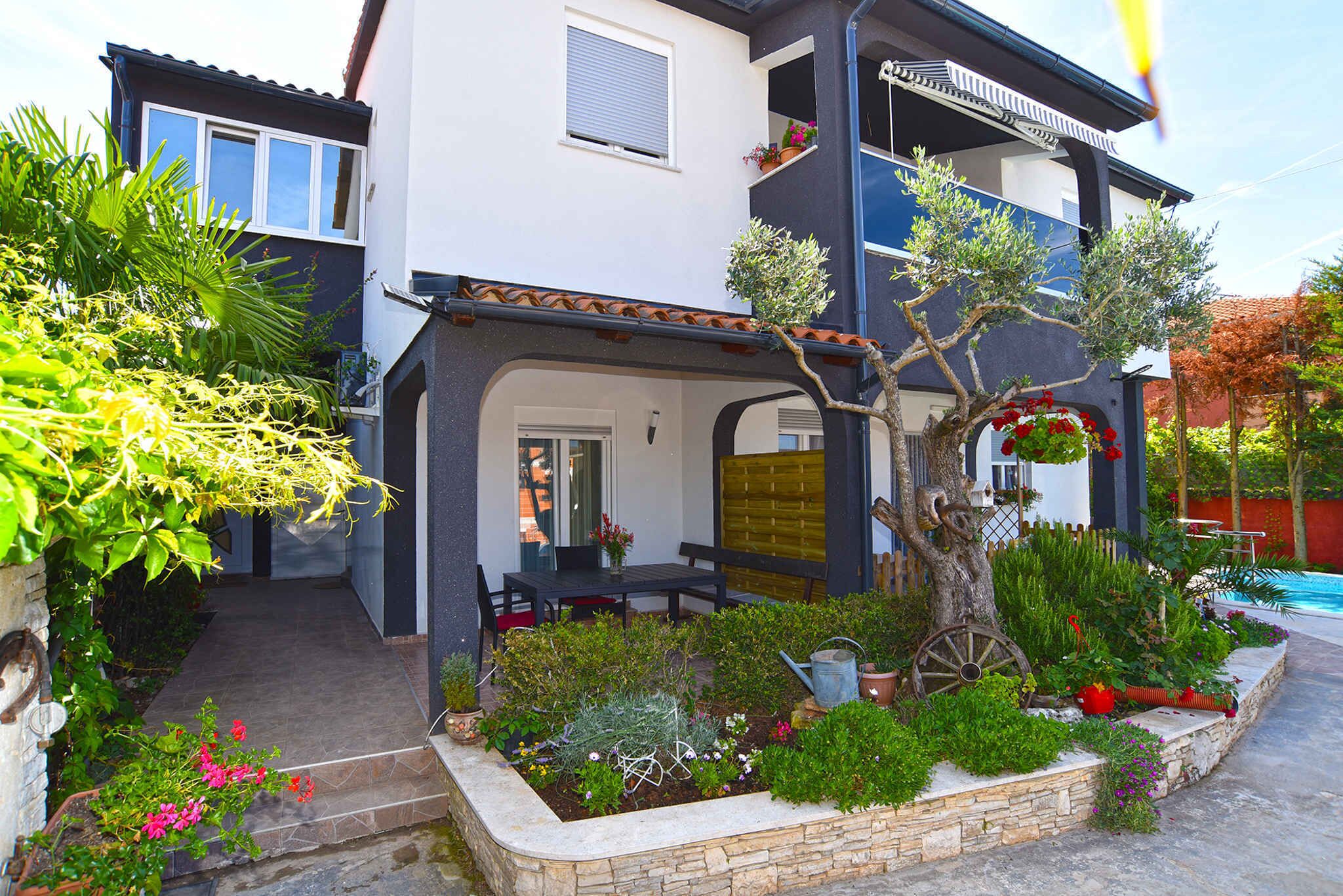 A two-story house with a balcony, a garden with various plants and flowers, and a small pool in the background.