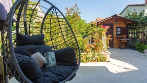 Ein Hängekorbstuhl mit Kissen und einem Buch, eine Terrassenanlage mit Pflanzen und ein Gebäude mit Holzfassade.