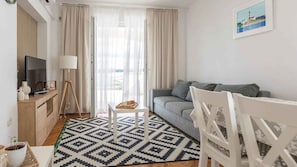 Un salon moderne avec un canapé gris, un fauteuil blanc, une petite table avec un plateau et un téléviseur posé sur un meuble en bois.
