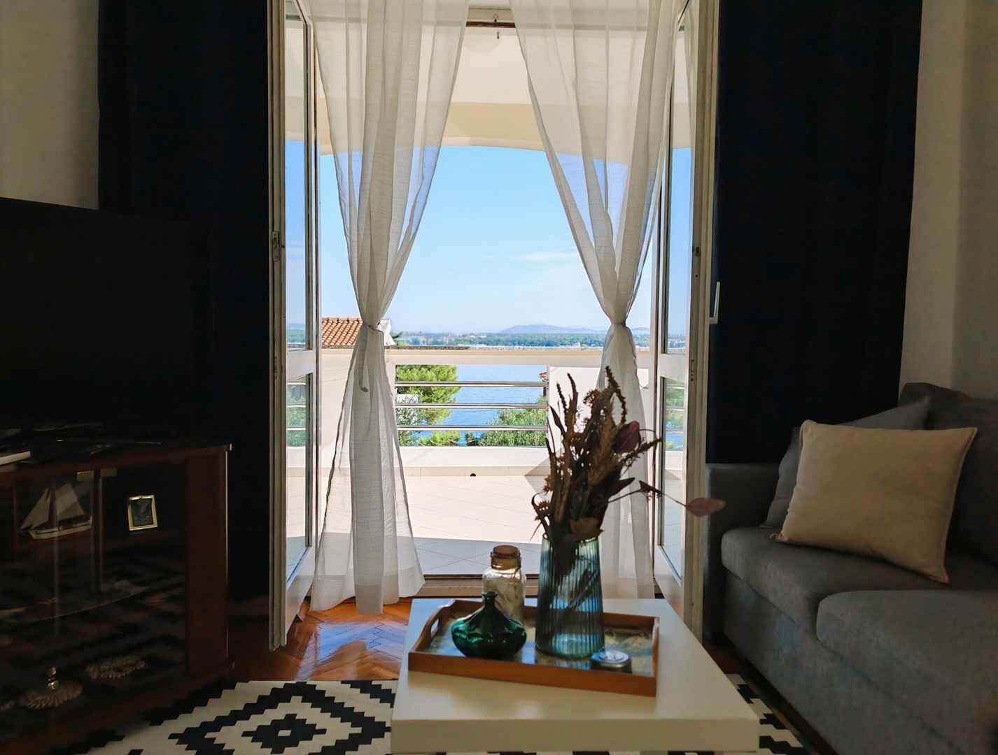 A living room with a sofa, a coffee table with a vase and a tray, and a view of a body of water through sheer curtains.