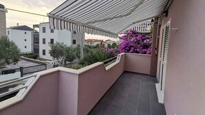 En balkon med en stribet markise, en blomsterkasse med lilla blomster og udsigt til omkringliggende bygninger.