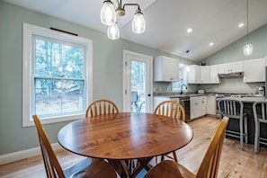 A kitchen with white cabinets, a wooden dining table, and a view of trees through large windows. - NEW!!! Charming 2-Bedroom in Beautiful Freeport! (Freeport)