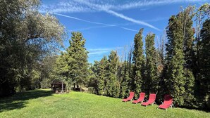 A grassy outdoor area with red lounge chairs and tall trees. - Holiday home with jetty and direct access to the water (Balatonmáriafürdő)