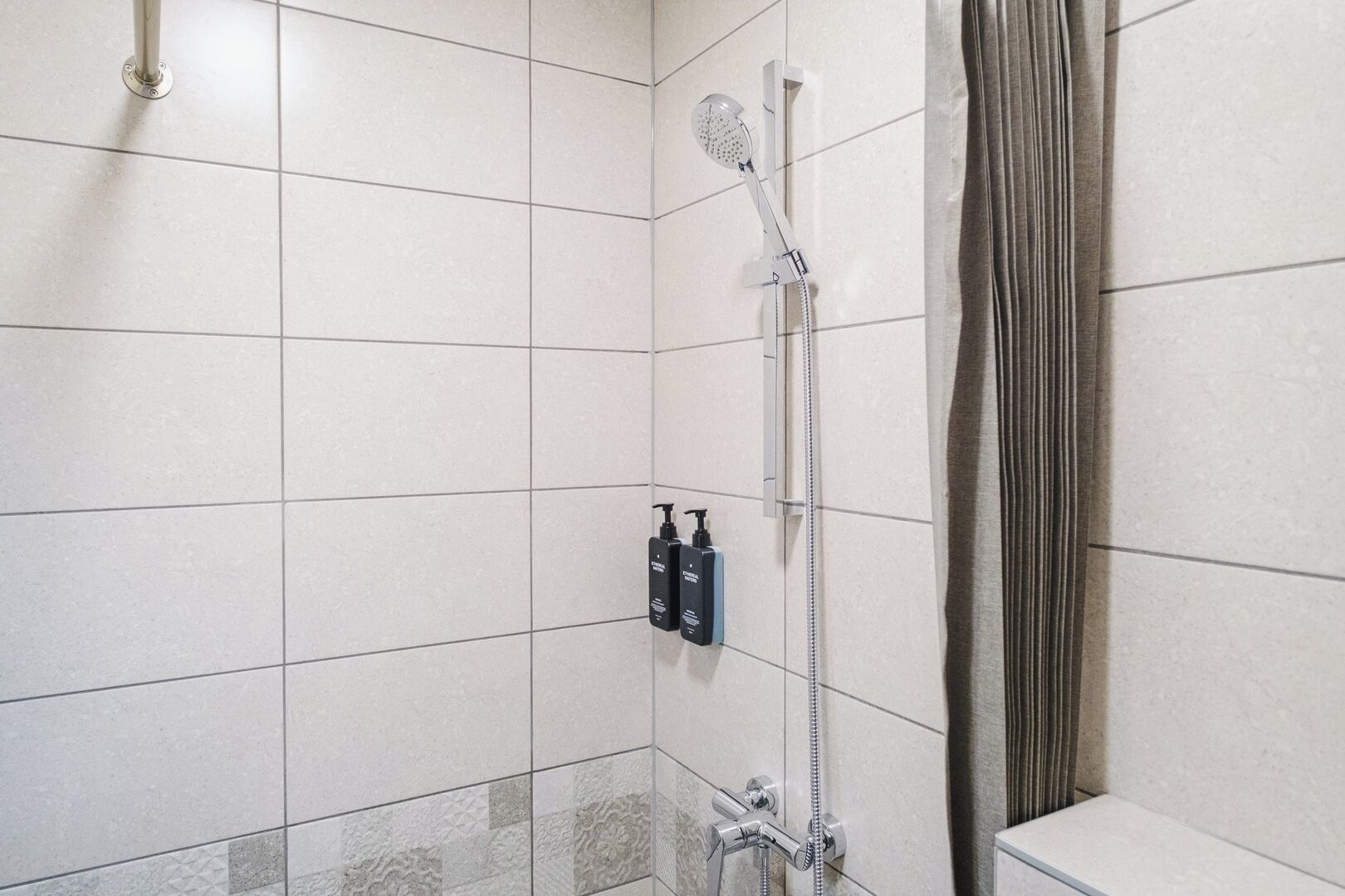 Bruseområde med håndbruser og vandhane på væggen. To shampoo- og balsambotler er monteret på væggen.