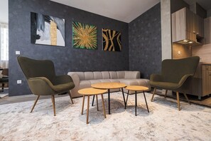 A modern living room with a sofa, two armchairs, and a coffee table. There are abstract paintings on the wall and a kitchen area to the right. - А17 Plovdiv Apartments Next to RowingChannel app17 (Plovdiv)