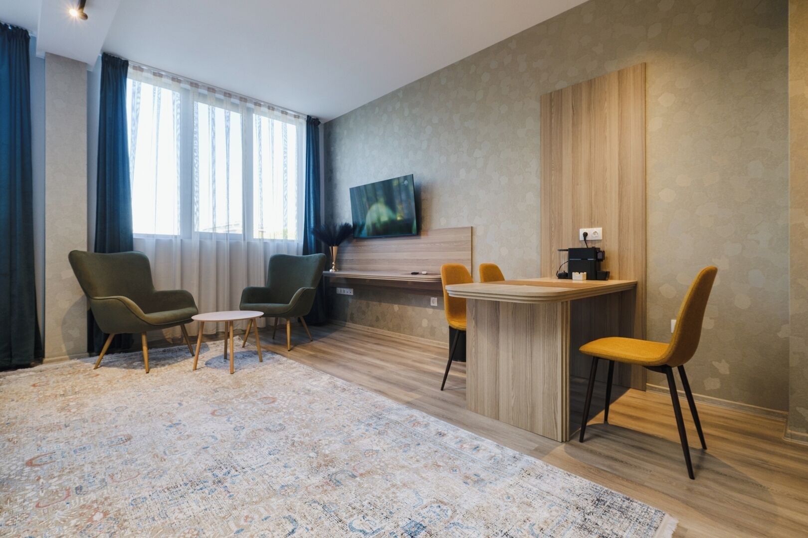 Een moderne woonkamer met een groot raam, een flatscreen televisie aan de muur, een houten eettafel met vier stoelen en twee fauteuils.