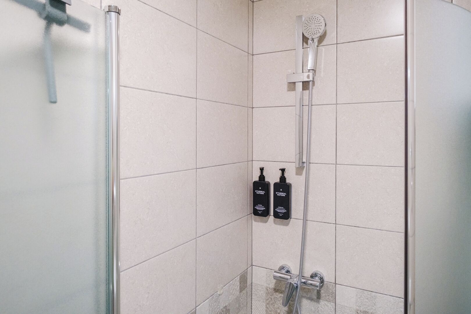 A shower area with a glass door, a handheld showerhead, and two bottles of Ethereal Waters on the wall.