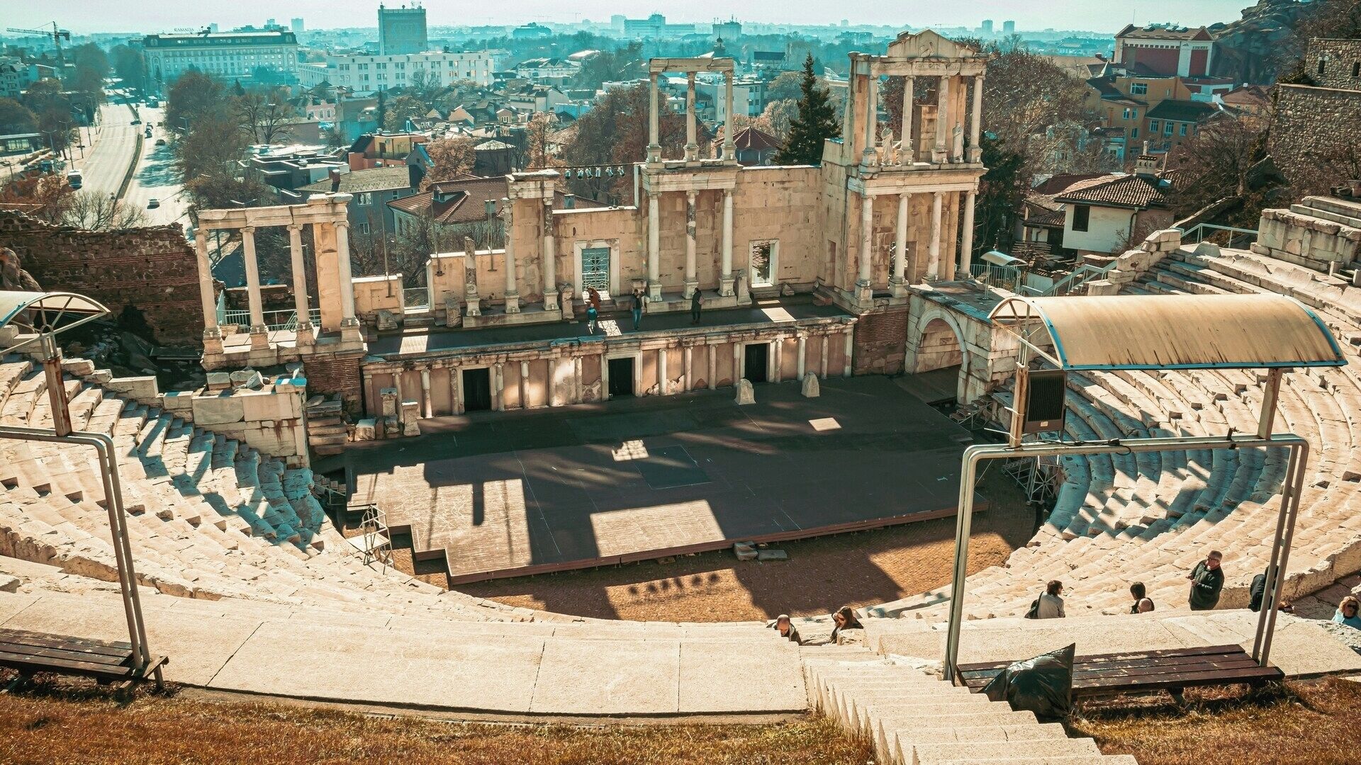An ancient amphitheater with stone steps and a central stage, surrounded by ruins and modern buildings.