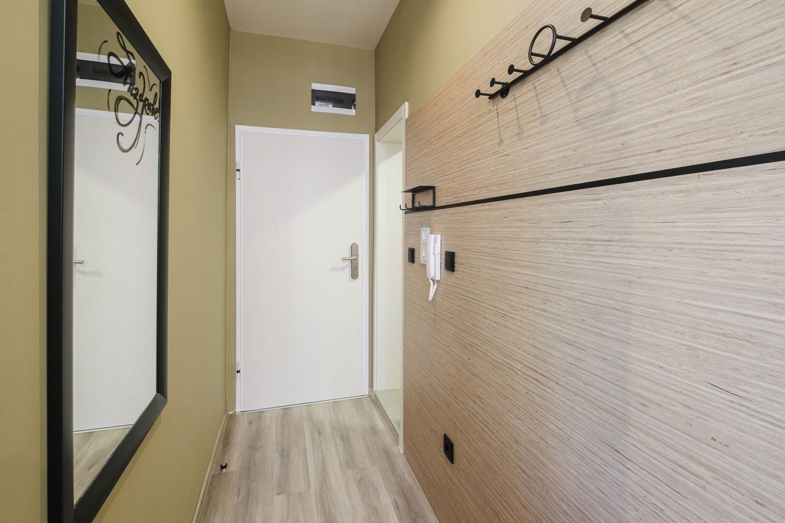A hallway with a wooden floor, a white door, a wall-mounted telephone, and a mirror with a decorative frame.