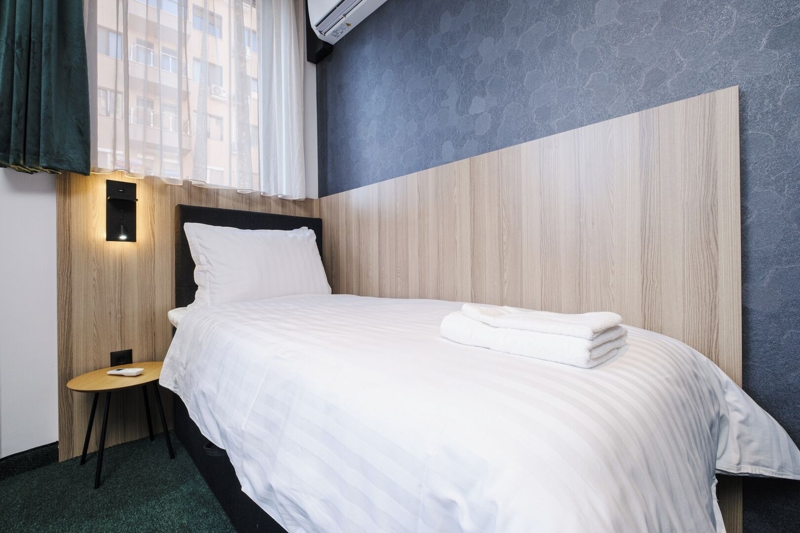 Una cama bien tendida con sábanas blancas y una toalla doblada, una pequeña mesa redonda con una lámpara, y un diseño moderno de pared con texturas de madera y piedra.