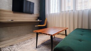 Una habitación moderna con un escritorio de madera, un sofá verde, un televisor montado y una ventana con cortinas translúcidas.
