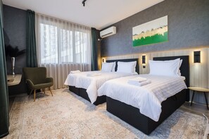 A hotel room with two beds, a desk, a chair, a large window with sheer curtains, an air conditioning unit, and a painting on the wall. - А17 Plovdiv Apartments Next to RowingChannel app16 (Plovdiv)