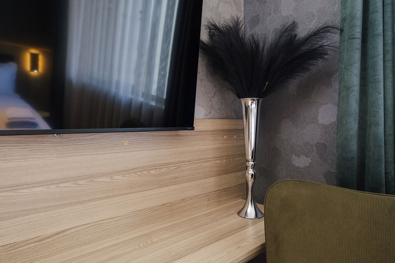 Een houten tv-meubel met een flatscreen televisie, een hoge metalen vaas met zwarte veertjes en een groen gordijn.