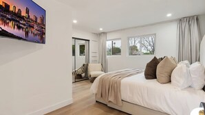 Une chambre moderne avec un grand lit, un téléviseur diffusant des images de la ville, et une vue sur l’extérieur par la fenêtre.