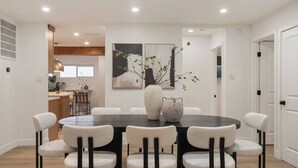 Un coin repas avec une table ronde, six chaises et deux vases. Au fond, on aperçoit une cuisine avec une fenêtre et une plante.