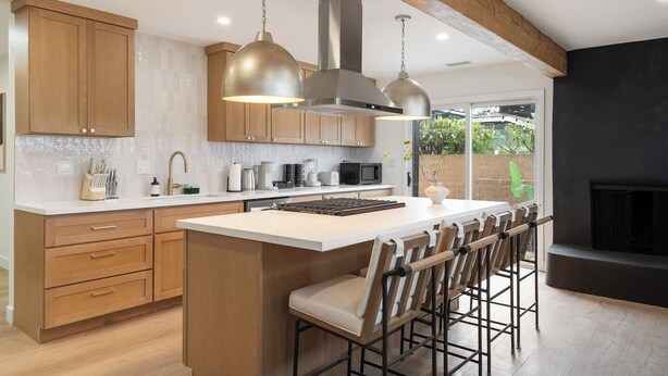 Une cuisine moderne avec des meubles en bois, un îlot central doté de tabourets de bar, un éclairage suspendu et une vue sur l’extérieur.