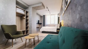 A modern hotel room with a green sofa, a wooden chair, a small round table, and a bed with a white duvet.