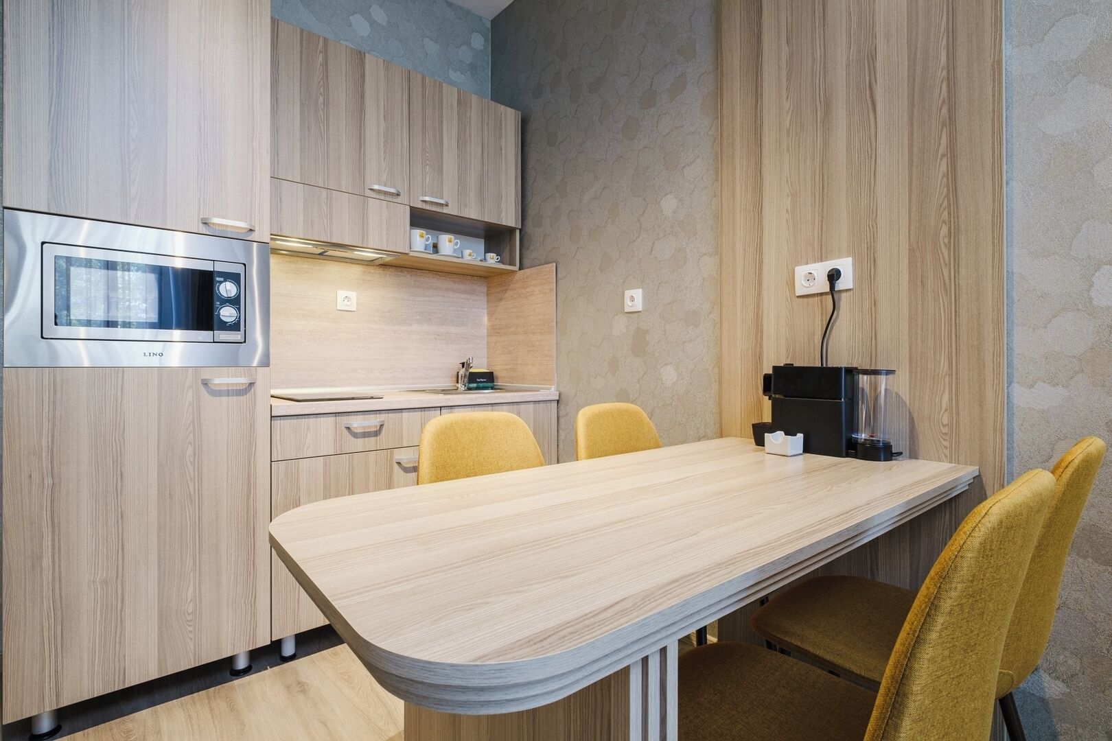 A modern kitchen with wooden cabinets, a built-in microwave, and a dining area with a curved table and yellow chairs.