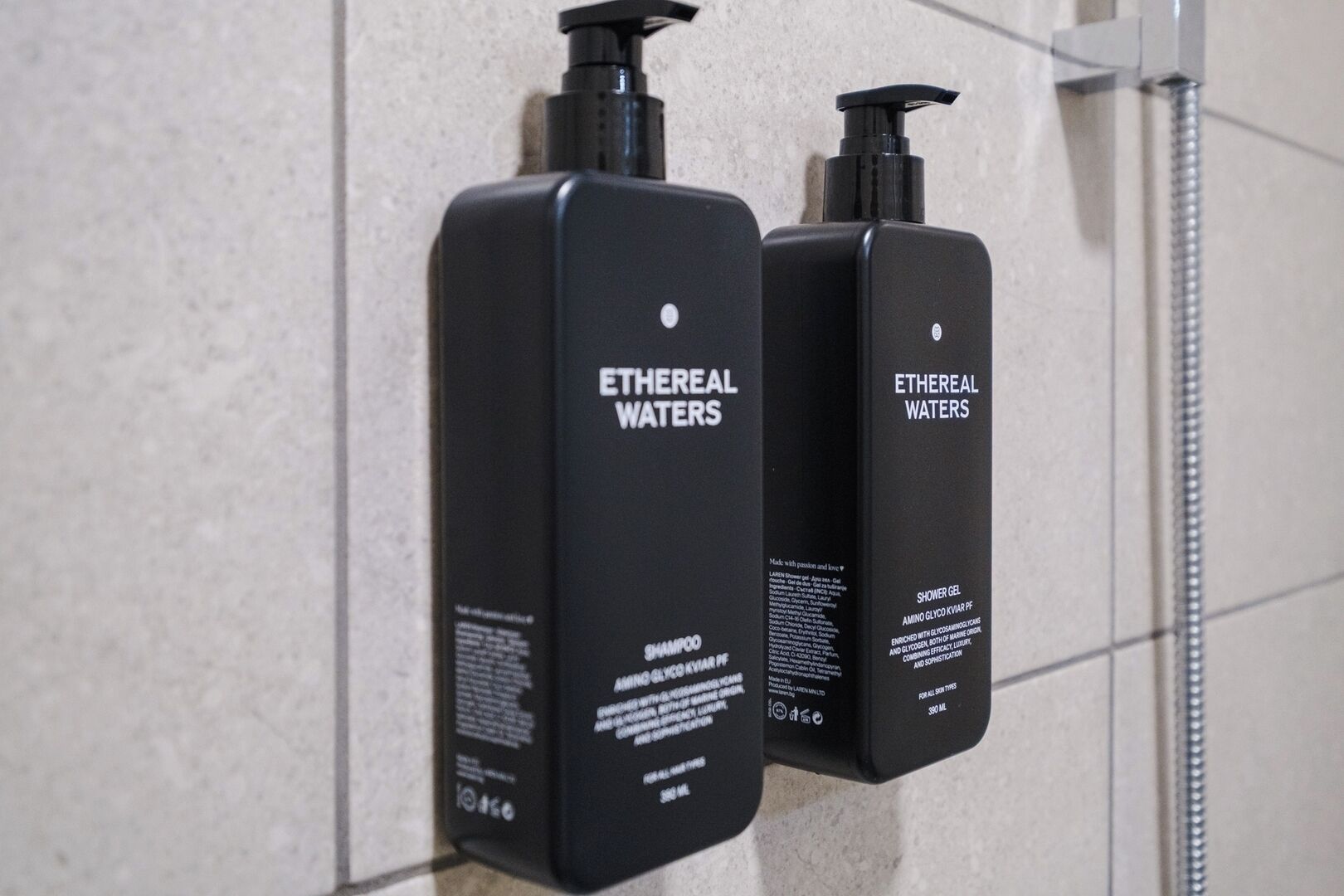 Deux flacons noirs de shampoing et de gel douche Ethereal Waters, chacun doté d’un distributeur à pompe, sont fixés à un mur carrelé.