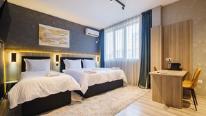 Une chambre d’hôtel avec deux lits, un bureau et une chaise. Il y a une grande fenêtre avec des rideaux, un climatiseur et un tableau au mur.