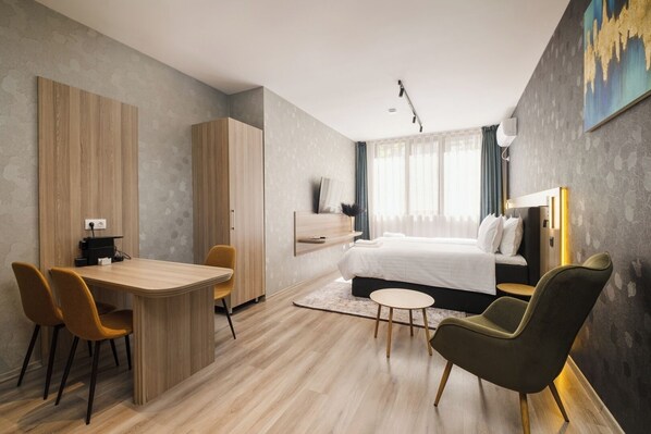 A modern hotel room with a wooden dining table, two chairs, a bed with white linens, a small round table, and a green armchair. - А17 Plovdiv Apartments Next to RowingChannel app7 (Plovdiv)