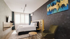 A modern hotel room with a large bed, a green armchair, a small round table, and a painting on the wall.