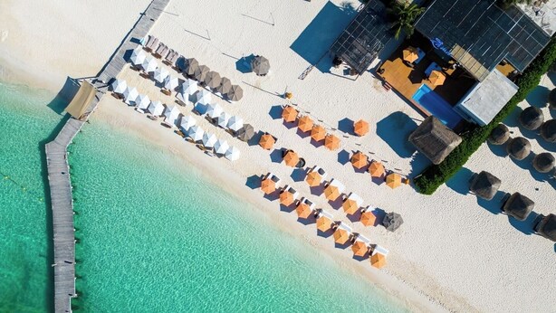 An aerial view of a beach with a wooden pier, rows of beach umbrellas, and a pool area. - Ocean Heaven: New Downtown Condo (Isla Mujeres)