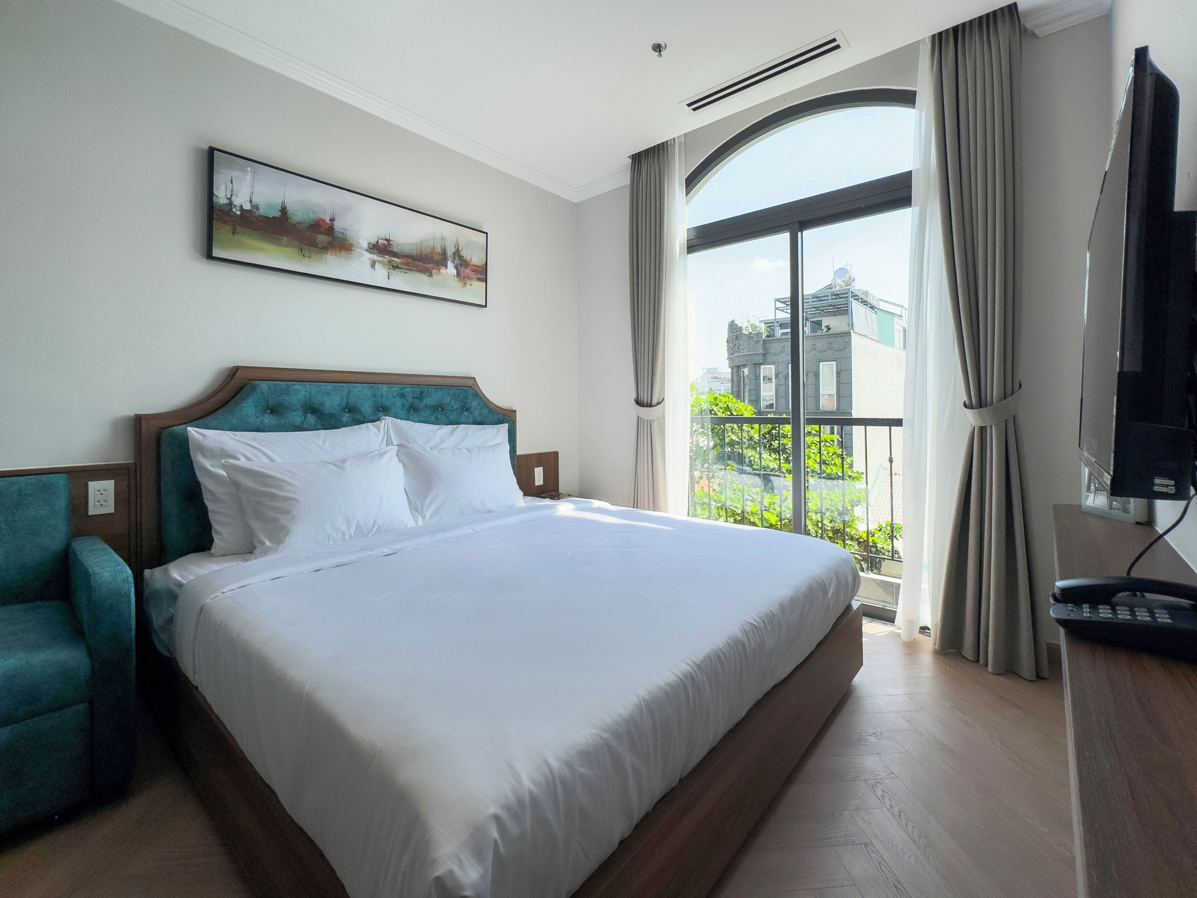 A hotel room with a large bed, a teal armchair, a painting on the wall, a TV, and a window with a view of buildings.