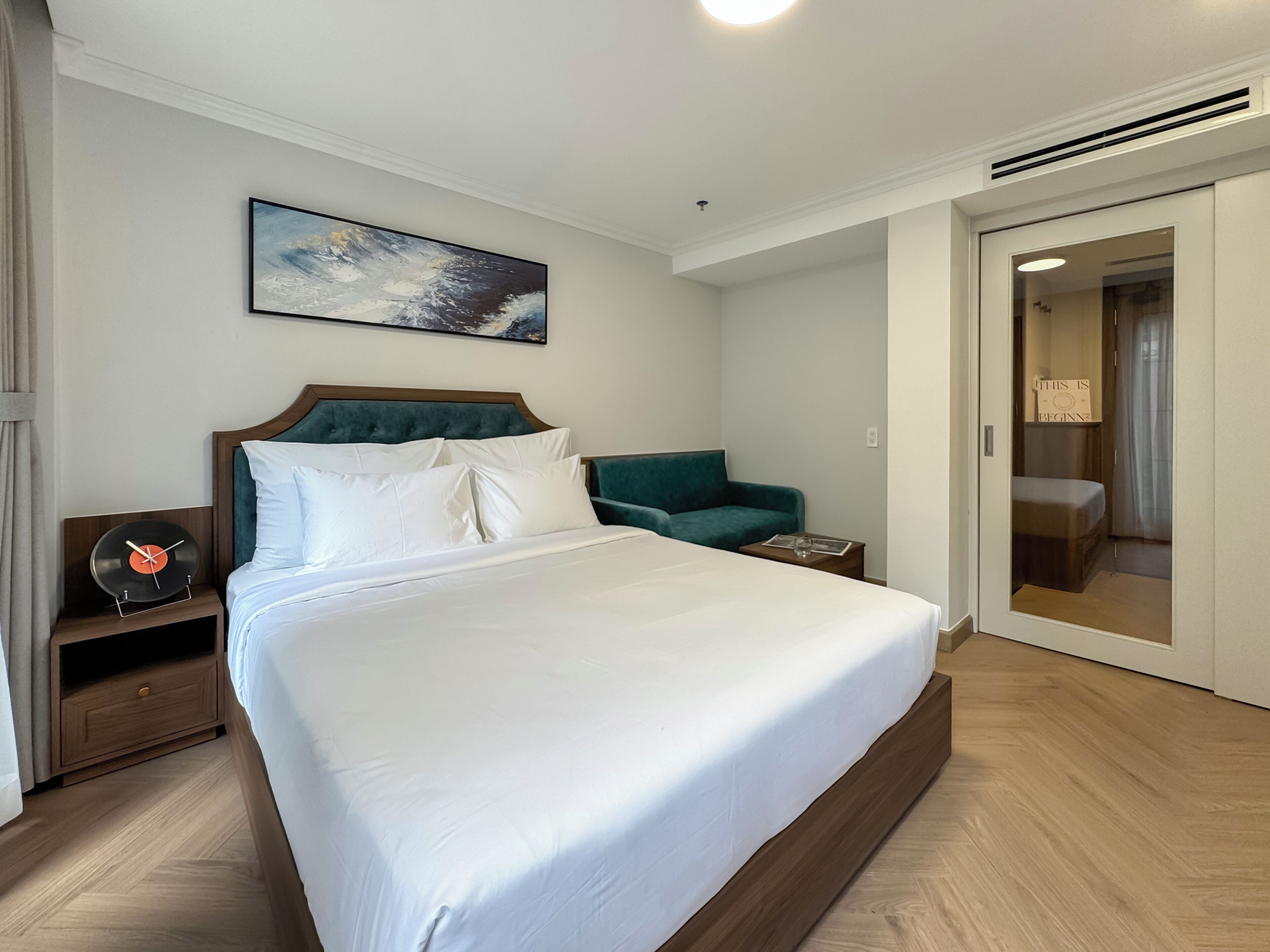 Habitación de hotel con una cama grande, un sillón verde, una mesita de noche con un tocadiscos y un cuadro en la pared.