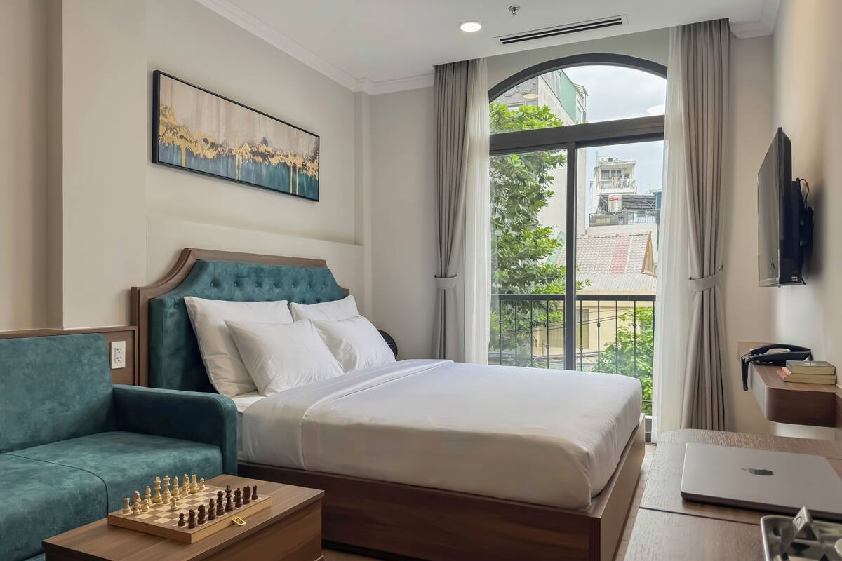 Ein Hotelzimmer mit einem Bett, einem Schreibtisch mit Laptop, einem Sessel, einem kleinen Tisch mit Schachbrett und einem großen Fenster mit Blick auf Gebäude und Grünflächen.