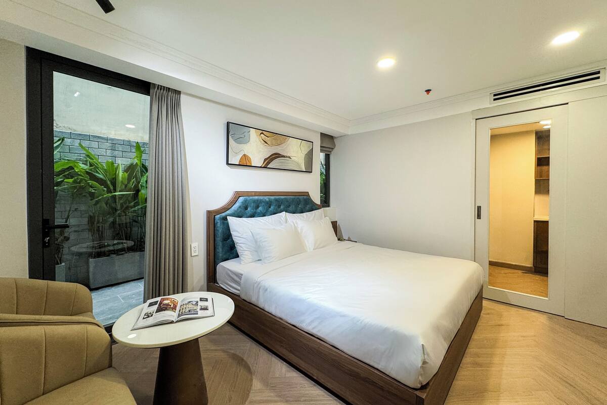 Ein modernes Hotelzimmer mit einem großen Bett, einem kleinen runden Tisch, einer Couch und einer Glastür zum Balkon mit Pflanzen.