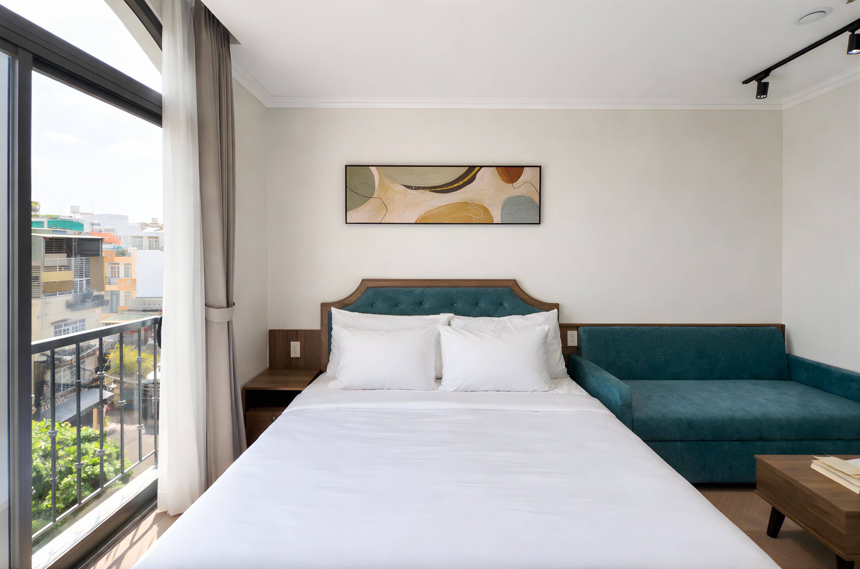 A hotel room with a large bed, a teal sofa, and a city view through a window.